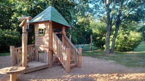 Spielplatz mit Bäumen und Holzhütte, die viel Schatten bieten