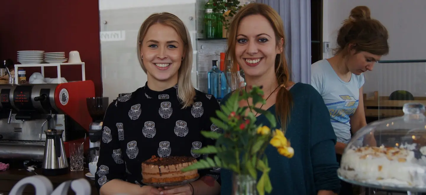 Zwei junge blonde Frauen schauen nebeneinander in die Kamera, im Hintergrund ein Café-Interieur.