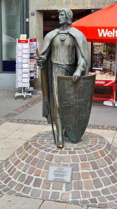 Man blickt auf eine Statue eines Mannes mit Schwert und Schild. Die Statue steht auf einem Sockel aus Pflastersteinen.