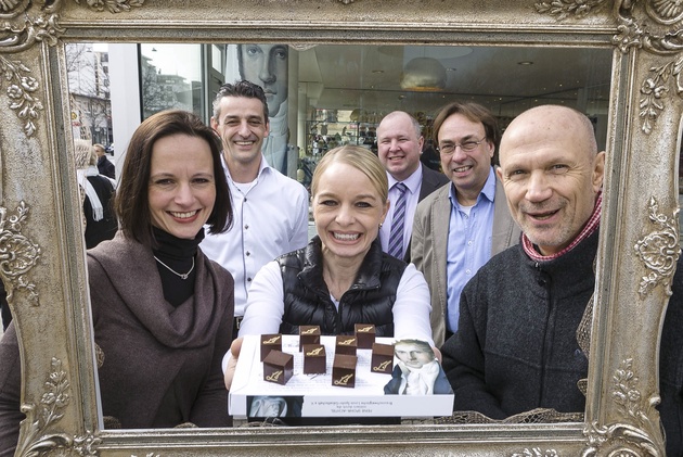 Eine Konditorin präsentiert ein Stück Konfekt. Im Hintergrund mehrere Leute.