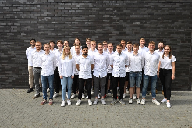 Eine Gruppe von zwanzig Studenten in weißen T-Shirts steht vor einer grauen Mauerwand.