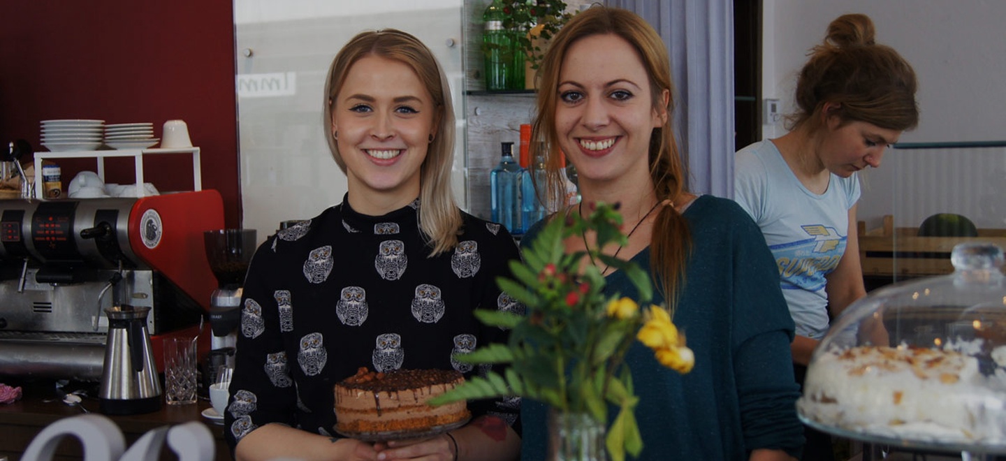 Zwei junge blonde Frauen schauen nebeneinander in die Kamera, im Hintergrund ein Café-Interieur.