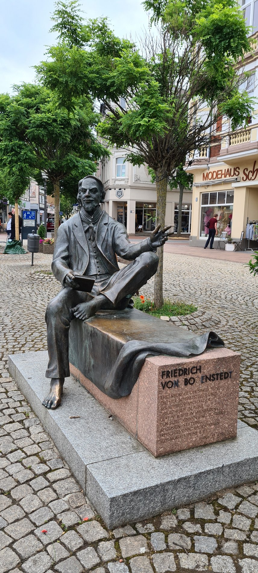 Man blickt auf eine Bronzestatue eines Mannes. Er sitzt auf einem Sockel aus grauem Granit und rotem Stein.