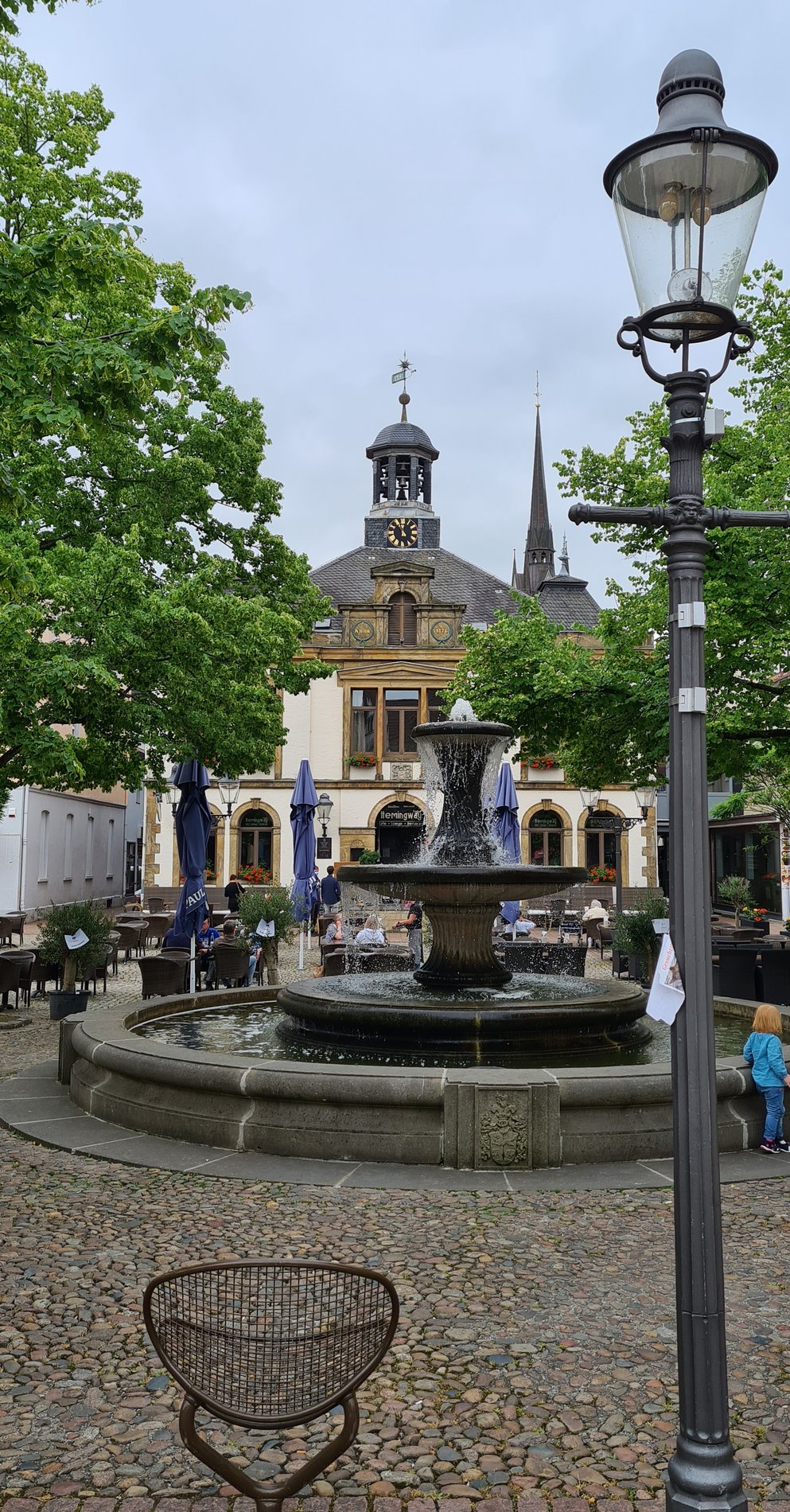 Man blickt auf einen geplasterten Platz in Mitten der Innenstadt. Auf dem Platz steht ein Springbrunnen. Im Hintergrund ist das alte Rathaus zu erkennen.