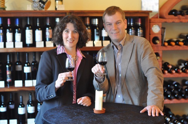 Ein Ehepaar mit jeweils einem Glas Wein in der Hand, vor dem Hintergrund eines Weinregals. 