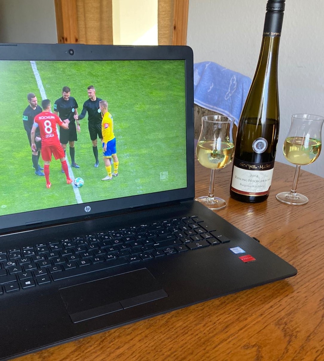 Ein Laptop zeigt eine Fußball-Übertragung, rechts daneben eine Flasche Weißwein mit zwei halbvollen Gläsern. 