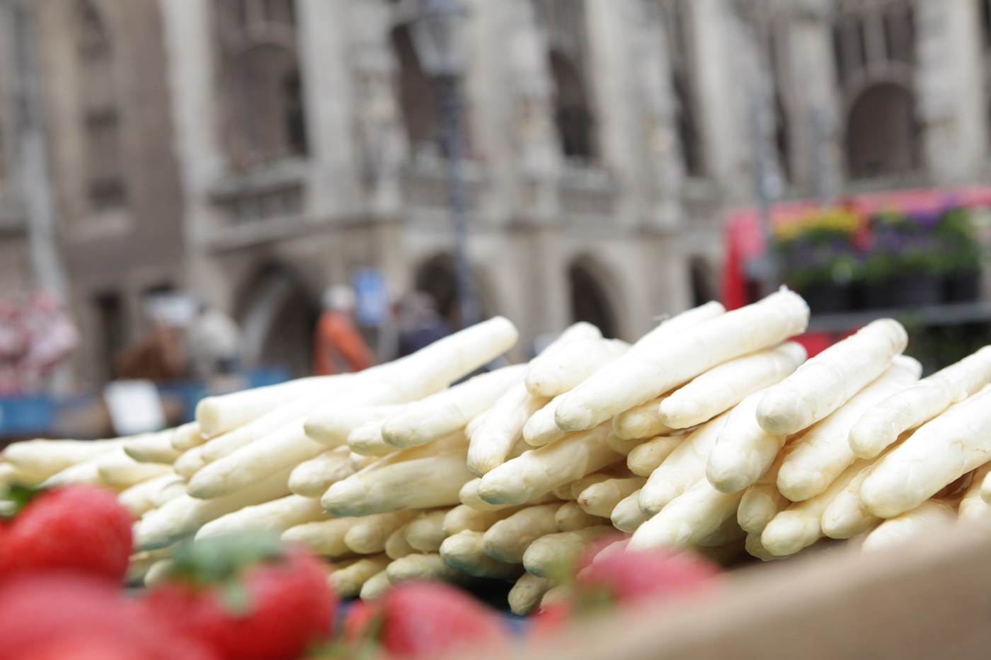 Auf dem Foto ist ein Haufen Spargel abgebildet. Im Hintergrund ist unscharf ein Sakralbau zu erahnen. Im Vordergrund, ebenso unscharf, sind einige Erdbeeren abgebildet.