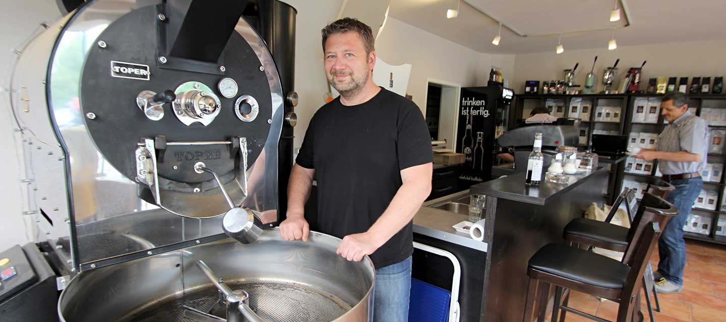 Ein Mann in schwarzem T-Shirt steht vor einem offenen Röstkessel für Kaffeebohnen.