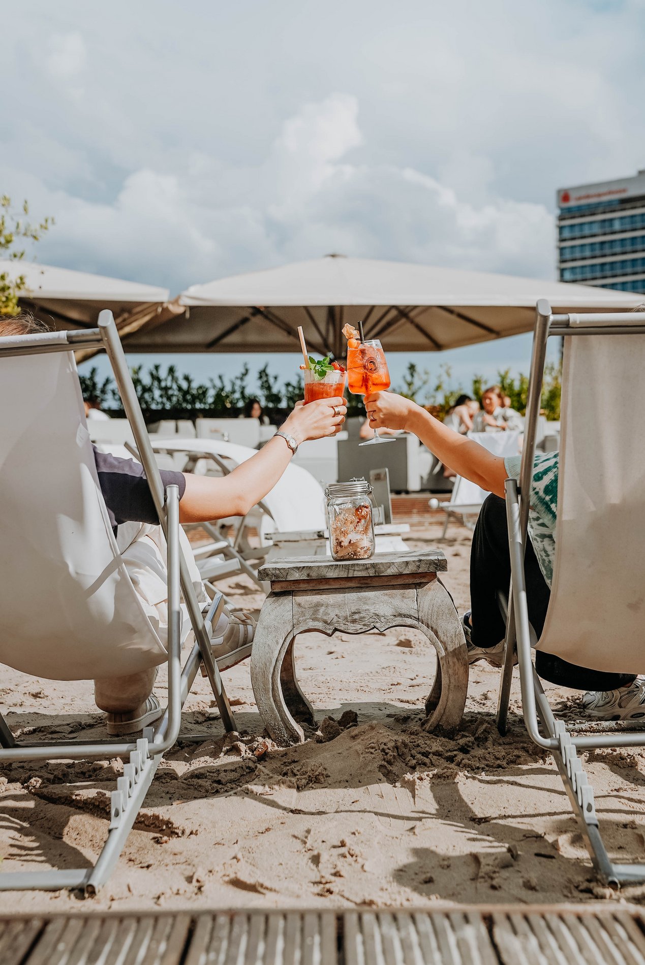 Zwei Menschen auf Liegestühlen in einer Beachbar, die mit einem Cocktail anstoßen 