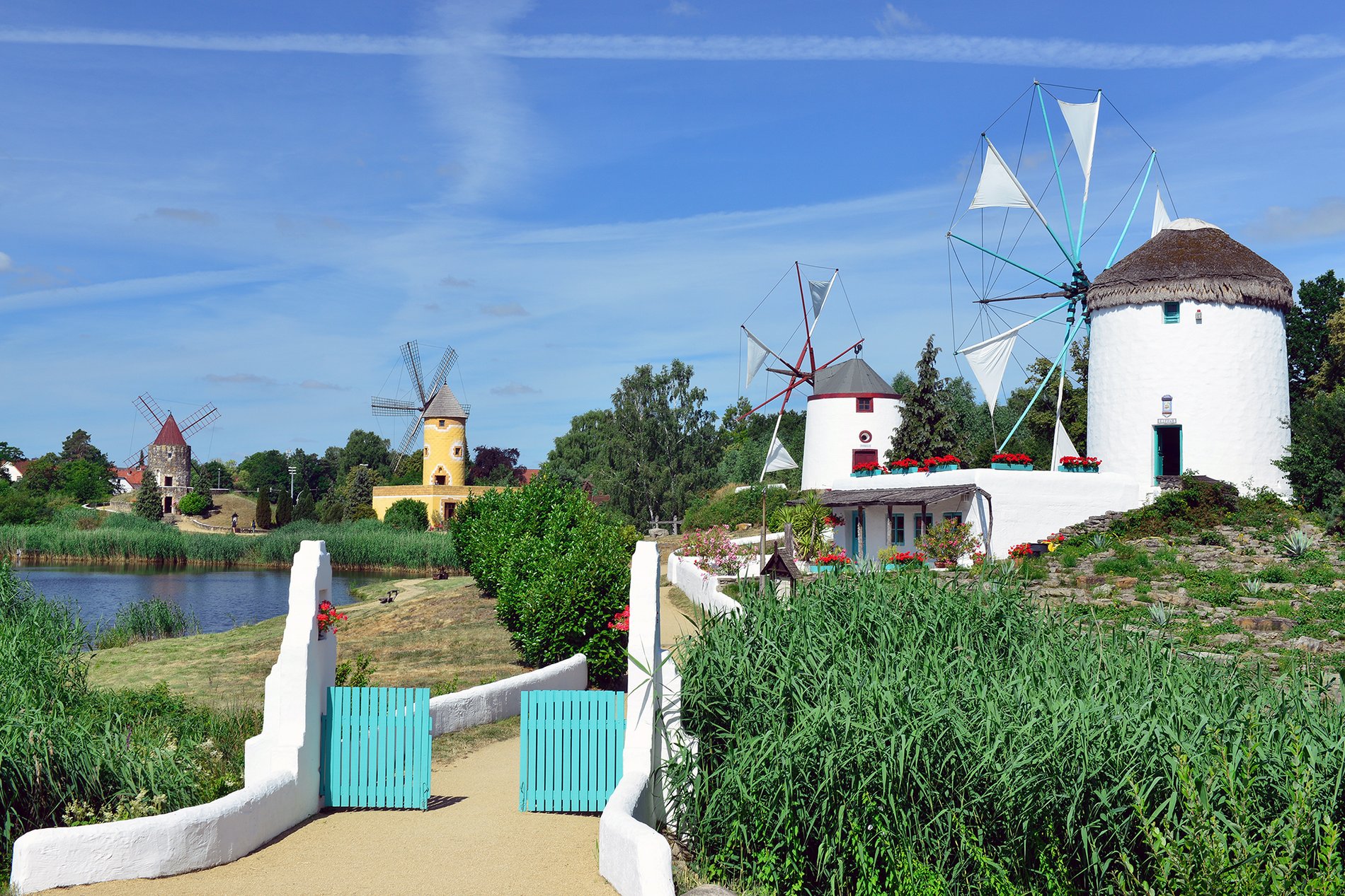 An einem Teich stehen viel Mühlen, umgeben von Sträuchern und Bäumen. 